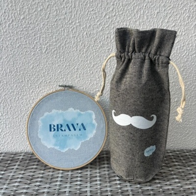 Saco de tecido cinzento com desenho de bigode branco e bastidor de bordado azul com texto BRAVA ESTAMPAGEM