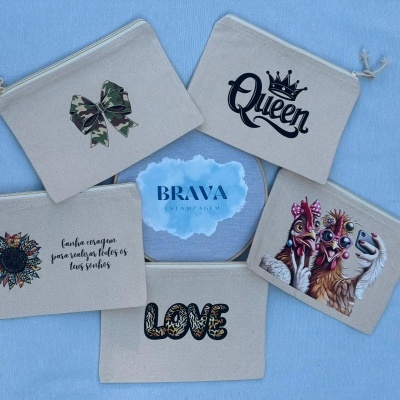 Cinco bolsas de tecido bege com diferentes desenhos e textos, dispostas sobre tecido azul.