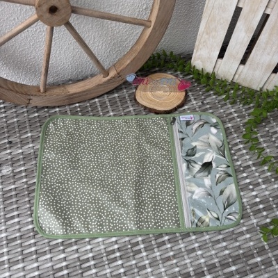 Carteira verde com padrão de bolinhas brancas e compartimento floral numa superfície de ráfia cinza com elementos de madeira e plantas