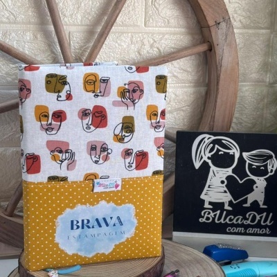 Capa de tecido colorida com rostos estilizados e bolinhas, em exposição sobre base de madeira, canetas e quadro decorativo próximo.