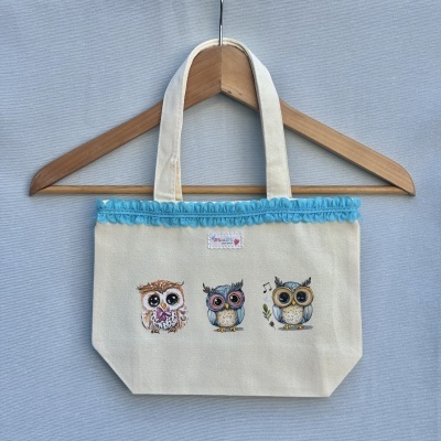 Saco de tecido bege com três corujas ilustradas e detalhe azul pendurado num cabide de madeira.