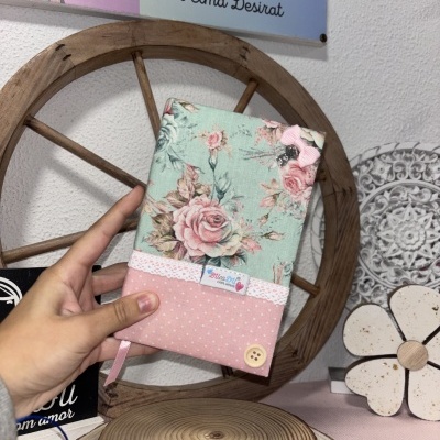 Capa de tecido floral e rosa com detalhes de renda e botão, segurada por mão em fundo rústico.