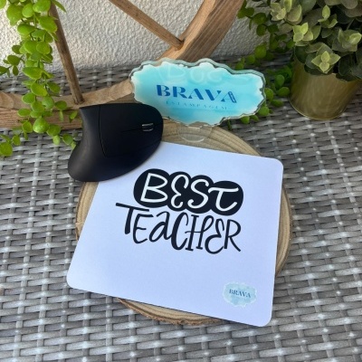 Tapete de rato branco com texto BEST TEACHER e selo azul sobre mesa com rato preto e plantas
