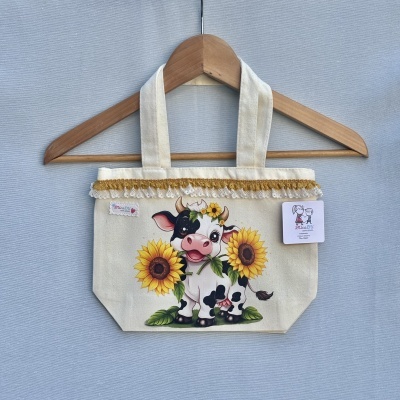 Saco de tecido cru com estampa de vaca e girassóis pendurado num cabide de madeira