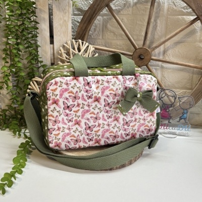 Mala retangular com padrão de borboletas, alças e laço verde, sobre base de madeira