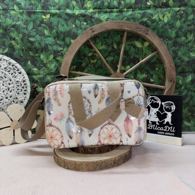Mala retangular creme com padrão de captores de sonhos e penas coloridas, com alças bege em tecido sobre suporte de madeira