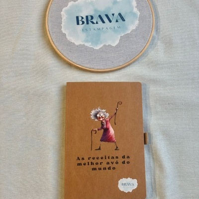 Caderno com capa castanha e ilustração de avó com cajado e bastidor de madeira com logo BRAVA ESTAMPAGEM.