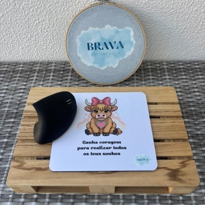 Mouse preto sobre tapete de rato com ilustração de vaca e texto motivacional em cima de suporte de madeira com bastidor ao fundo