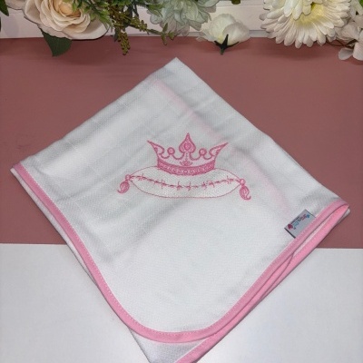Manta branca com borda rosa e bordado de coroa rosa, sobre superfície rosa e branca com flores artificiais ao fundo