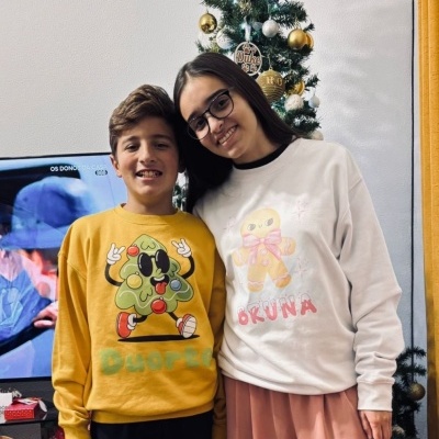 Dois jovens sorrindo com sweatshirts coloridas frente a árvore de Natal decorada.