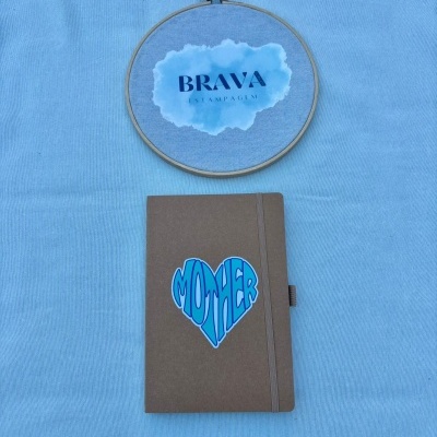 Caderno castanho com autocolante azul em coração MOTHER e aro de bordado com texto BRAVA ESTAMPAGEM