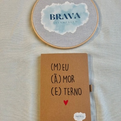 Caderno castanho com texto em português ao lado de bastidor de bordado com logotipo BRAVA