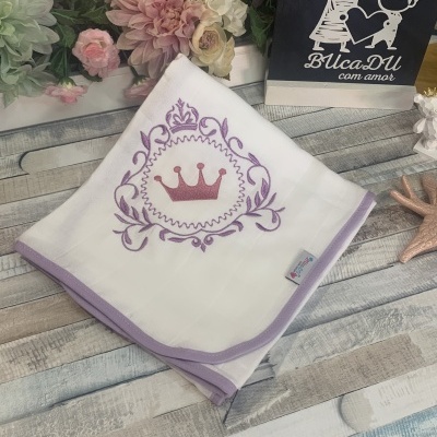 Manta branca com bordado roxo de coroa sobre superfície de madeira com flores artificiais e placa preta