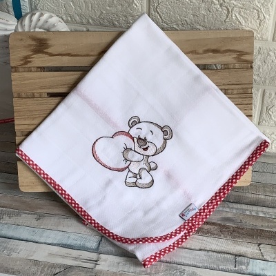 Fralda branca com borda vermelha e bordado de urso, sobre bancada de madeira.