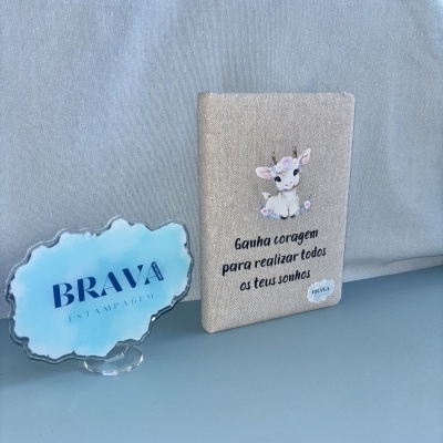 Caderno bege com cabrito e frase motivacional ao lado de expositor azul com logo BRAVA