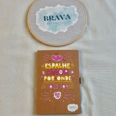 Caderno castanho com mensagem colorida e aro de madeira com texto BRAVA ESTAMPAGEM em pano cinzento