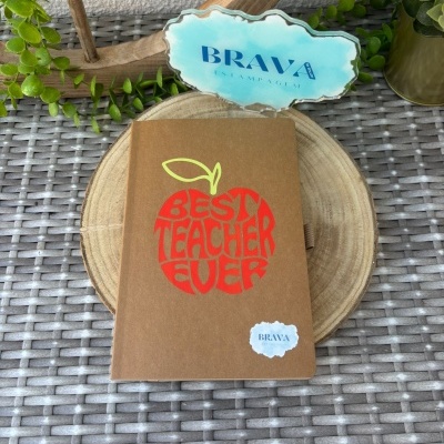 Caderno com capa castanha e maçã vermelha com texto, sobre disco de madeira e fundo com plantas.