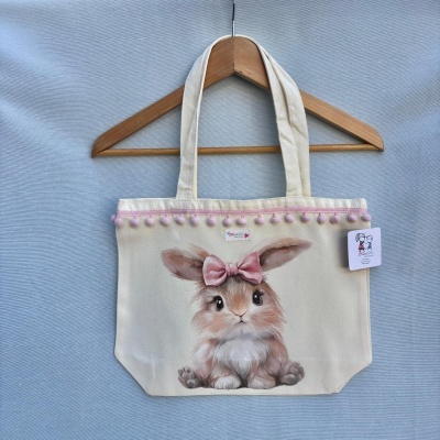 Saco de tecido creme decorado com coelho pintado e pompons cor-de-rosa pendurado num cabide de madeira