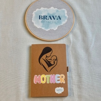 Caderno castanho com ilustração de mãe e bebé e texto MOTHER ao lado de bastidor com tecido e logo BRAVA ESTAMPAGEM
