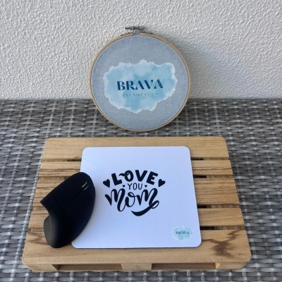Rato de computador e tapete de rato branco com texto LOVE YOU mom em base de madeira sobre superfície de rattan cinzento e bastidor de bordado com texto BRAVA
