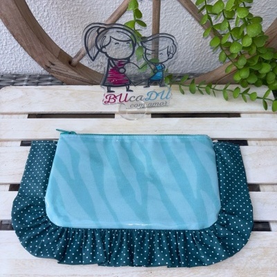 Necessaire azul claro com acabamento em tecido azul escuro com bolinhas brancas sobre uma superfície de madeira clara com decoração de folhagem verde e roda de madeira ao fundo