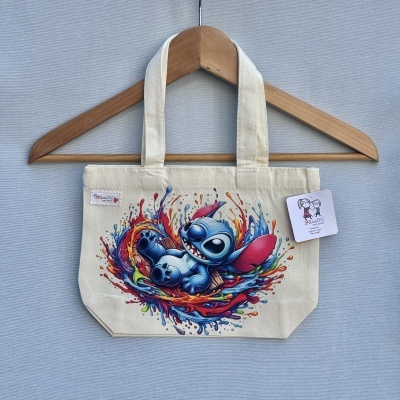 Sacola bege com estampa de personagem azul e respingos coloridos, pendurada em cabide de madeira