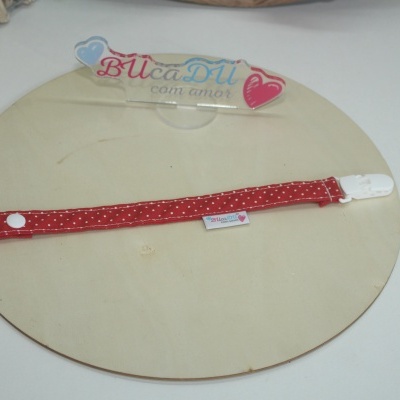 Presilha para chupeta vermelha com bolinhas brancas e fecho branco sobre fundo branco e madeira clara