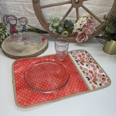 Jogo americano vermelho com bolinhas brancas e prato de vidro sobre ele, copo transparente e fundo decorativo de roda de madeira e flores.