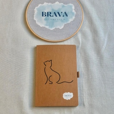 Caderno castanho com desenho de gato e aro de tecidos com texto BRAVA ESTAMPAGEM