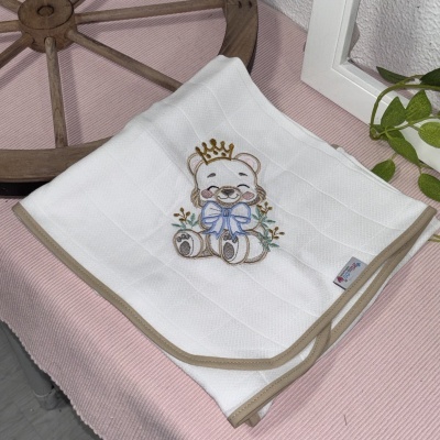 Toalha branca bordada com ursinho e coroa sobre tecido rosa com roda de madeira e plantas