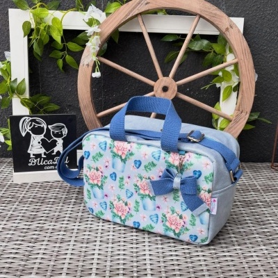 Mala pequena floral com alças azuis e laço, em superfície trançada, com roda de madeira e moldura branca com plantas ao fundo.