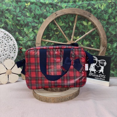 Mala xadrez vermelho com alças azuis e laço, em suporte de madeira com fundo de folhas verdes.