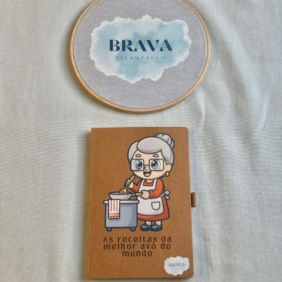 Caderno marrom com ilustração e texto As receitas da melhor avó do mundo e cómodo forrado a tecido cinza claro com logo BRAVA ESTAMPAGEM.