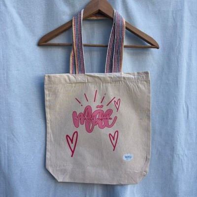 Tote bag de algodão cru com alças multicoloridas e texto vermelho sobre fundo azul claro.