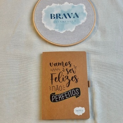 Caderno com capa castanha e frase motivacional ao lado de aro de bordado com logo BRAVA