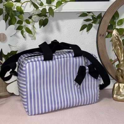 Mala de tecido às riscas lilás e branca com alças pretas sobre superfície rosa e placa decorativa atrás