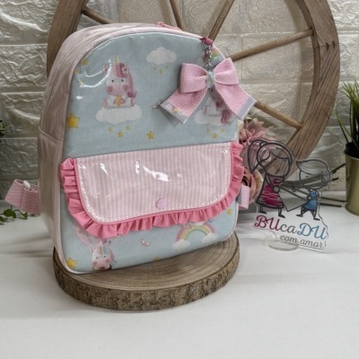 Mochila infantil azul e rosa com unicórnios, laço rosa e bolso frontal, sobre base de madeira