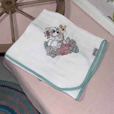 Manta branca bordada com coelho, flores e passarinho numa superfície rosa