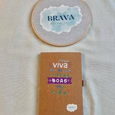 Caderno castanho com texto colorido e bastidor com tecido e logo BRAVA ESTAMPAGEM