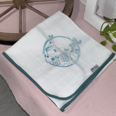 Pano de cozinha branco bordado com dinossauros azuis e planta, borda azul, sobre tecido rosa ao lado de roda de madeira e planta.