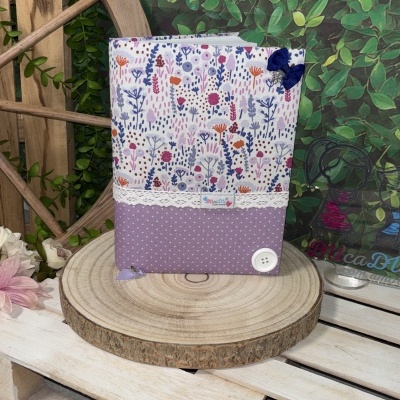 Capa de livro em tecido floral e lilás com botão e laço, sobre base de madeira