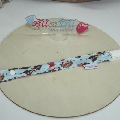 Pulseira em tecido azul claro com padrão de bonecas e corações, com fecho branco e etiqueta BUcaDU com amor sobre base redonda de madeira.