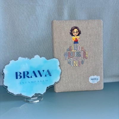 Caderno cinza com texto e ilustração de professora e peça acrílica azul com texto Brava Estampagem