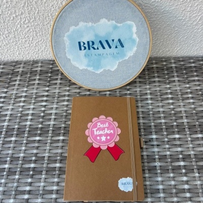 Caderno castanho com etiqueta Best Teacher sobre esteira cinza e aro de madeira com texto BRAVA ESTAMPAGEM