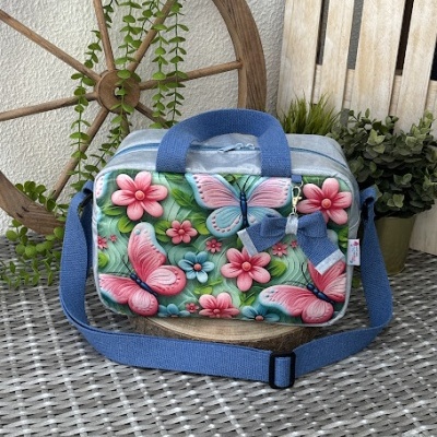 Mala pequena com borboletas e flores cor-de-rosa e azul, com alça azul, em ambiente com roda de madeira e plantas.