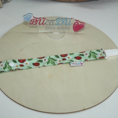 Porta-chupetas tecido verde com padrão joaninhas e flores, fechado branco, sobre madeira clara