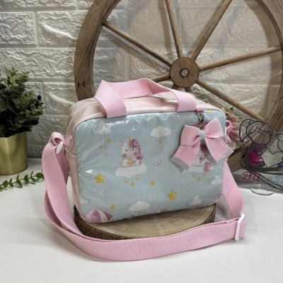 Bolsa infantil azul com padrão de unicórnios e estrelas, com alça rosa, sobre mesa branca e roda de madeira ao fundo
