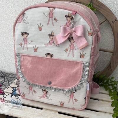 Mochila rosa clara com padrão de bonecas e coroas com bolso frontal e laço rosa.