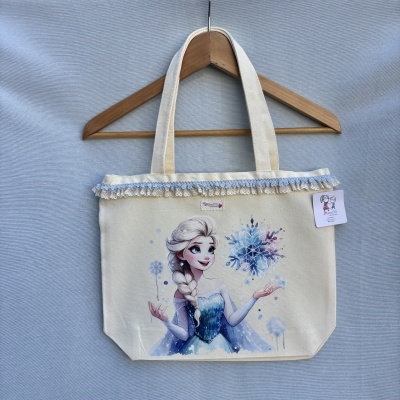 Saco de tecido creme com desenho da Elsa do Frozen e rendas azuis pendurado num cabide de madeira