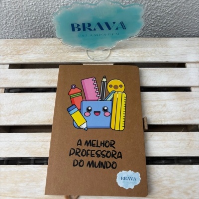 Caderno castanho com desenho de material escolar e texto A MELHOR PROFESSORA DO MUNDO encostado num suporte azul claro sobre madeira clara
